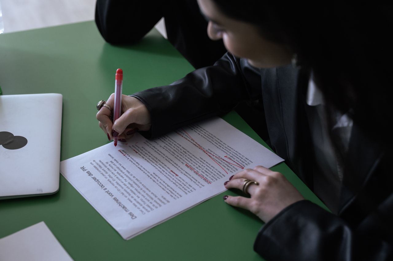 A woman in a suit uses a red pen to edit a printed document on a green table with a laptop